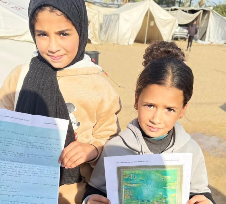 Two girls hold papers, standing in a camp setting with tents in the background.