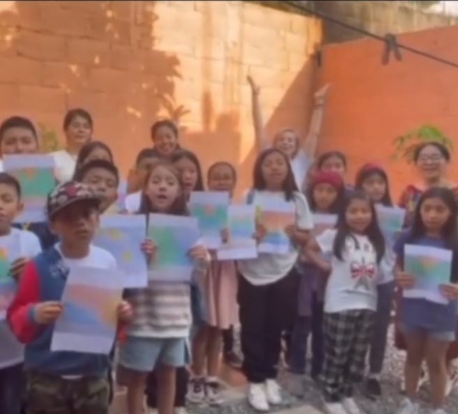 Group of children holding artwork, smiling and celebrating outdoors against an orange wall.