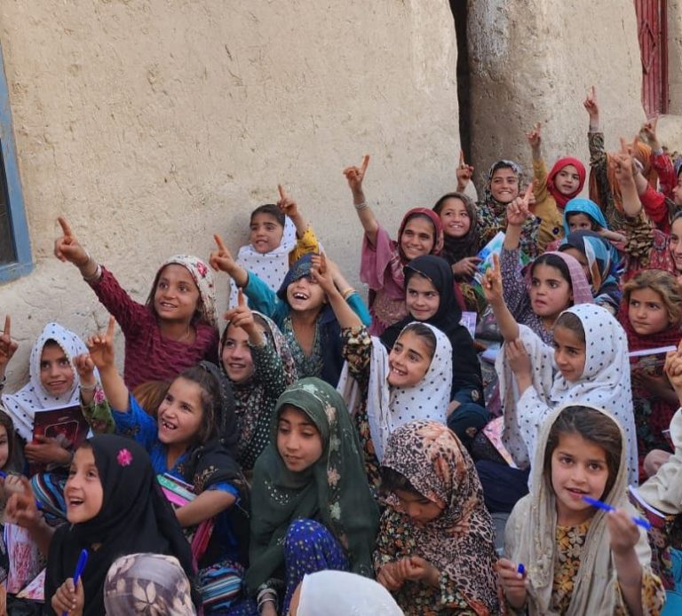 Group of children holding artwork, smiling and celebrating outdoors against an orange wall.