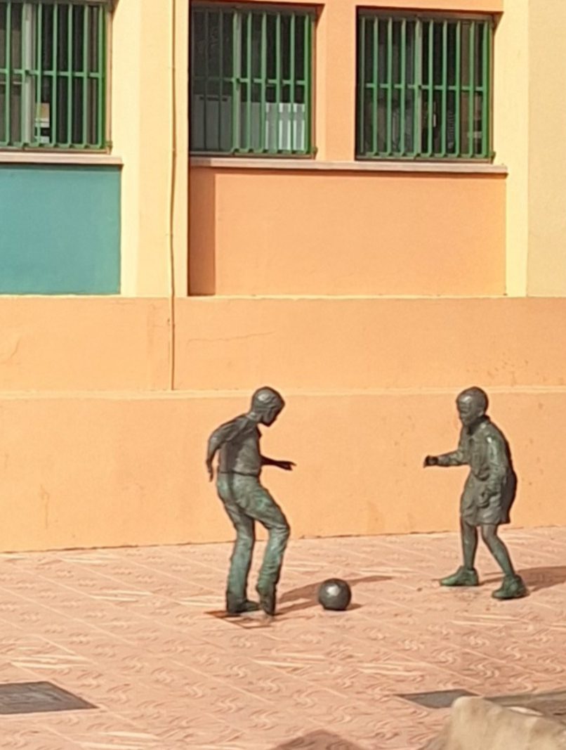 Two bronze sculptures of children playing football on a patterned pavement.